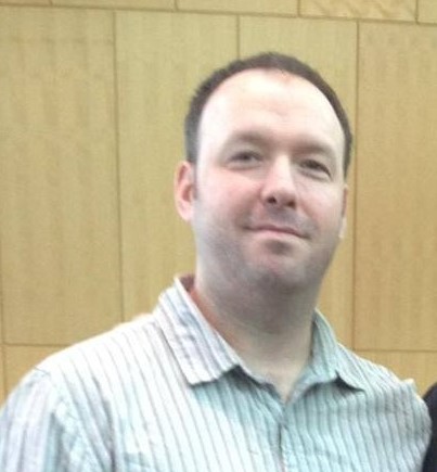 Daniel Canavin Photo Person in a striped shirt smiling, standing in front of a wooden panel background.